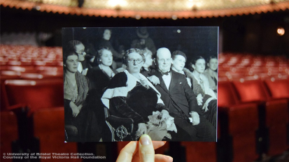 A person holds a black and white photography of Lilian Baylis seated in The Old Vic stalls, lining it up with the modern seats in the auditorium
