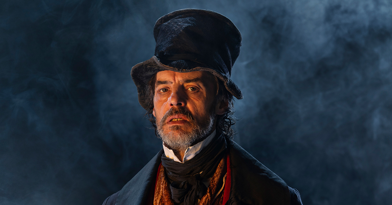Actor Paul Hilton glowers at you against a dark background with white smoke. He wears a crumpled top hat, a velvet reddish-brown waistcoat, a large overcoat and a stiff collar.