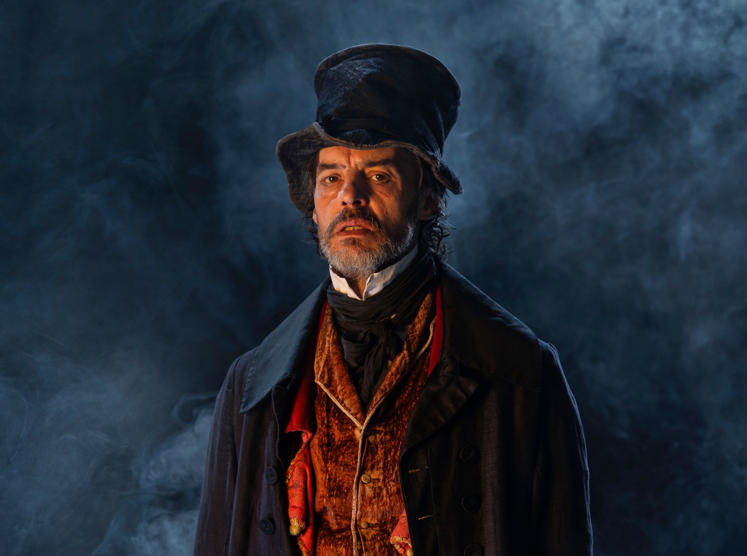 Actor Paul Hilton glowers at you against a dark background with white smoke. He wears a crumpled top hat, a velvet reddish-brown waistcoat, a large overcoat and a stiff collar.