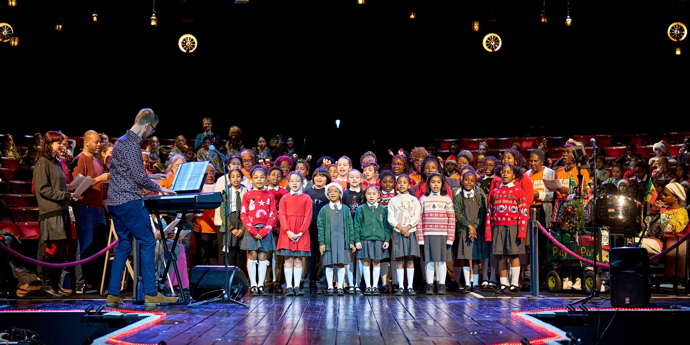 A group of school children and adults are on stage at The Old Vic singing as a choir and wearing Christmas accessories and jumpers.