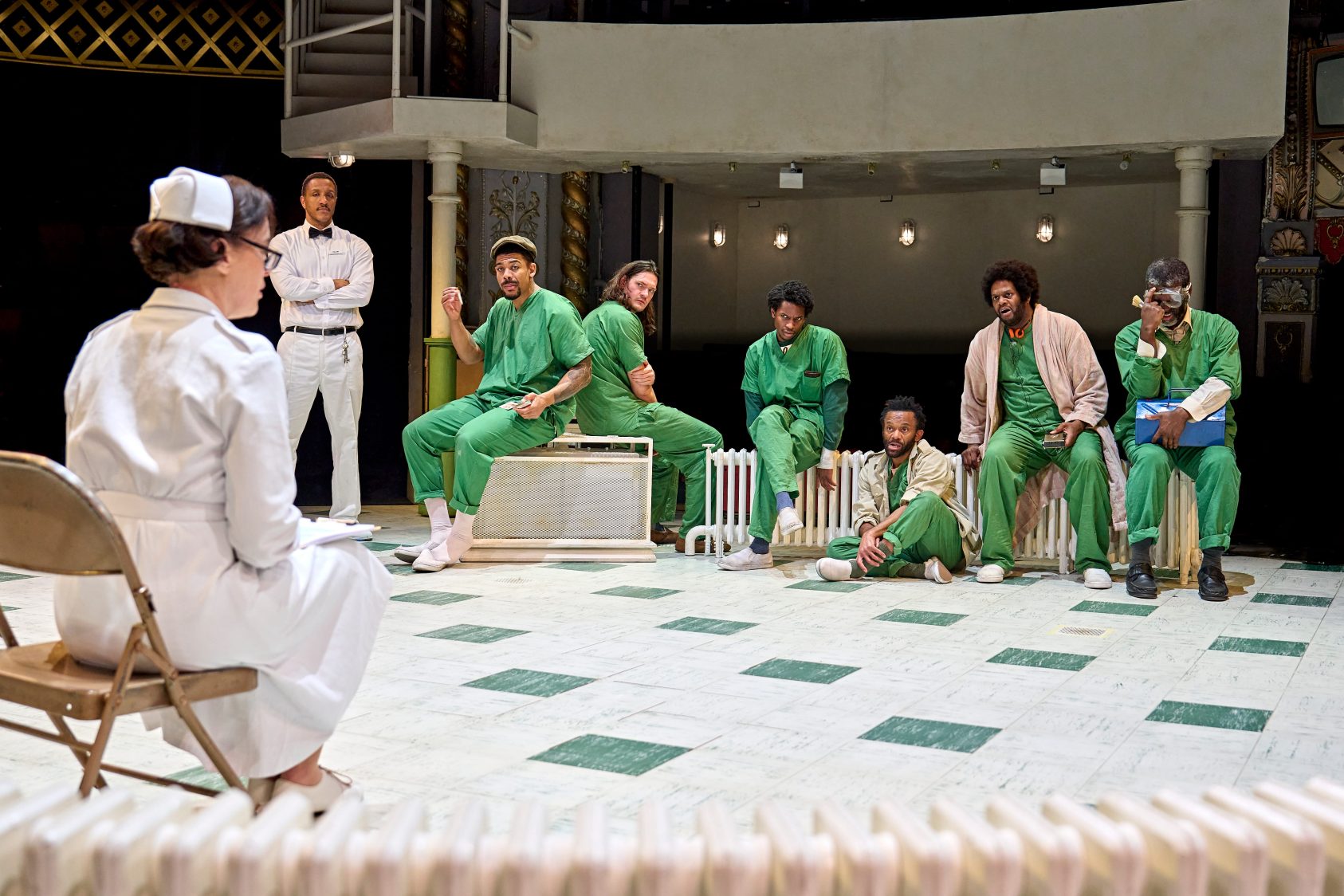 Actor Olivia Williams, wearing a white nurse's uniform with a white hat, sits on a folding chair with her back to us, she's looking at some of the cast of One Flew Over the Cuckoo's Nest, who are wearing green patient's outfits.