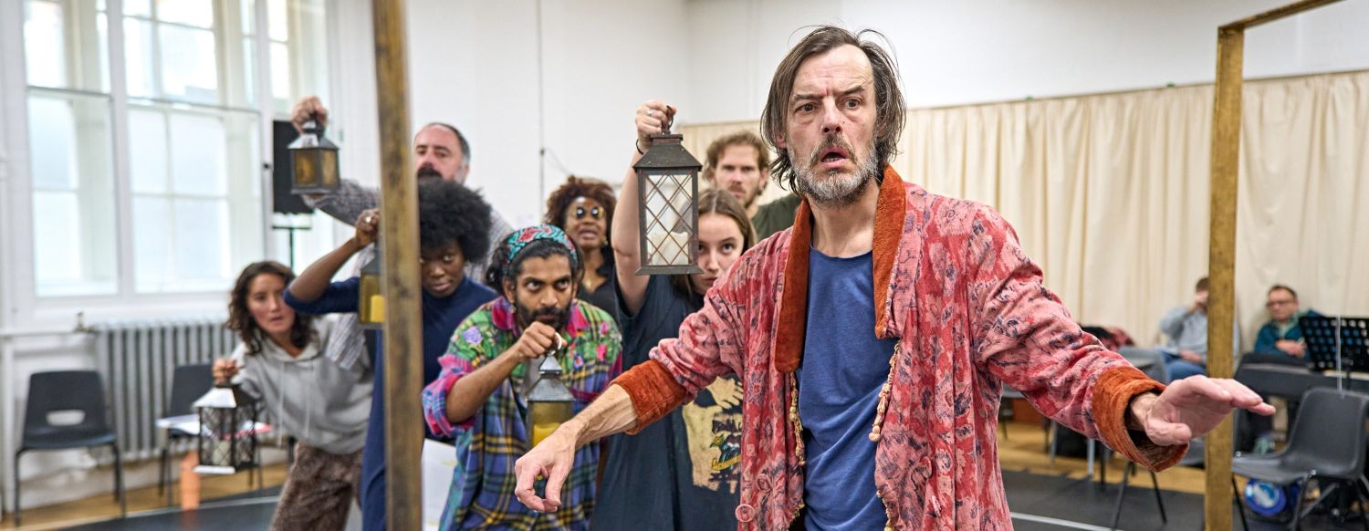 Paul Hilton stands in front of the cast of A Christmas Carol, wearing a red dressing gown over exercise clothing.