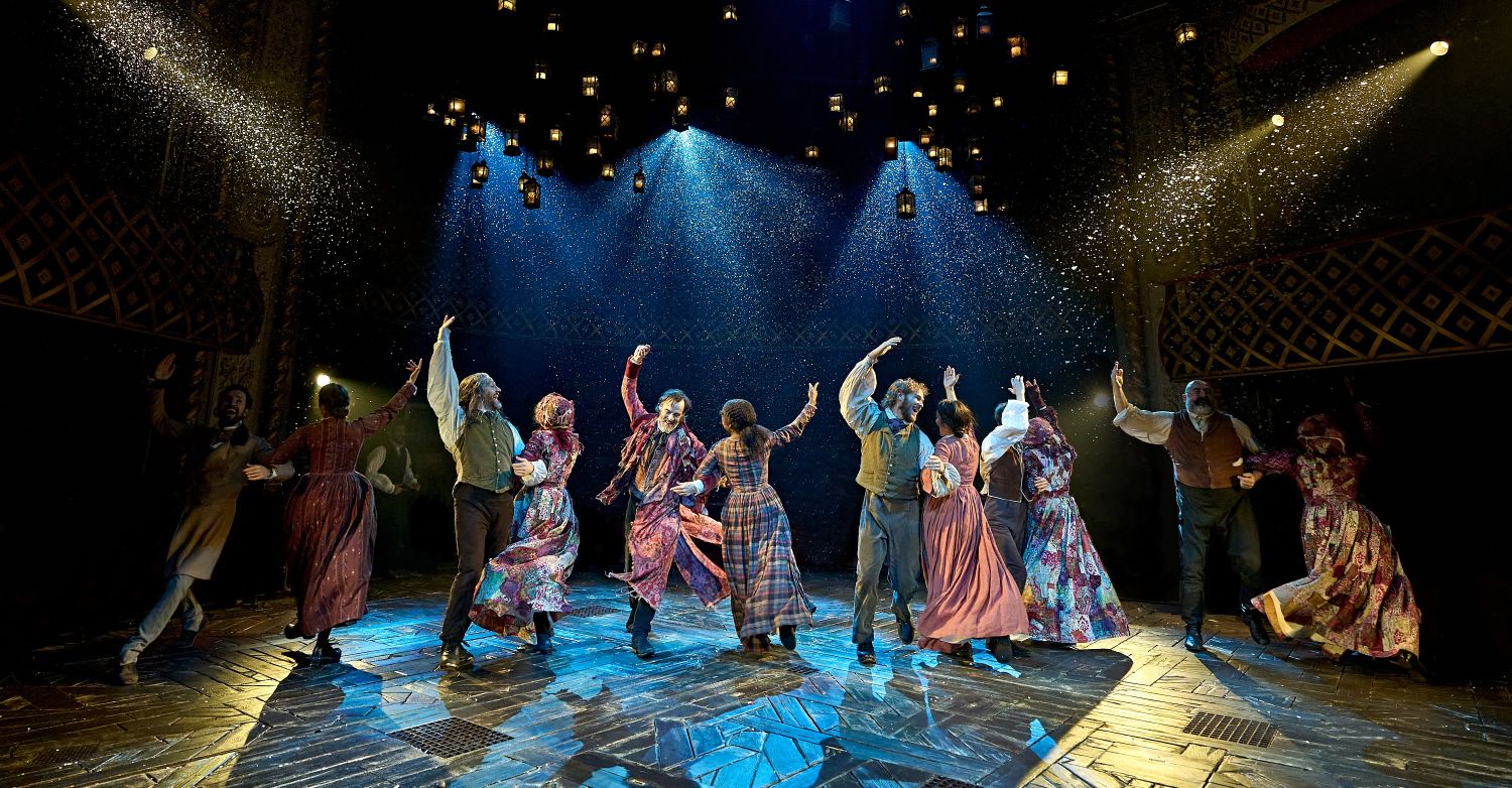 Six actors are dancing a jig in pairs, one arm interlinked with their partner and the other raised. Above them, snow falls down from cannons and tens of glowing lanterns are hanging from the rafters.