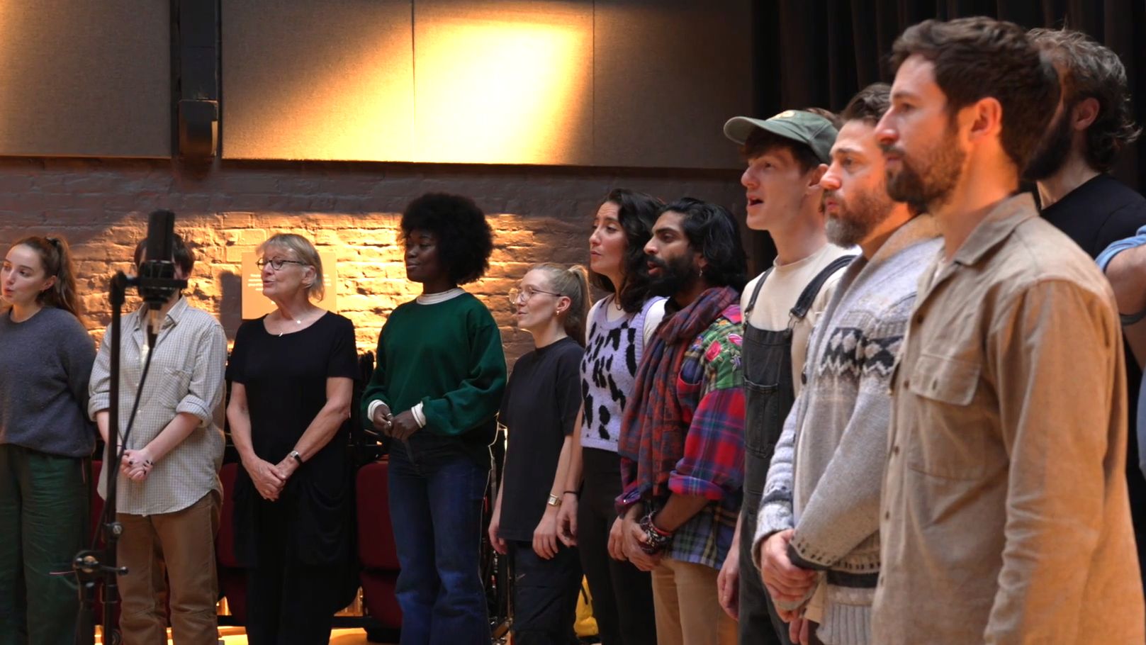 The cast of A Christmas Carol stand in a line in a rehearsal room, singing into a microphone on a stand.