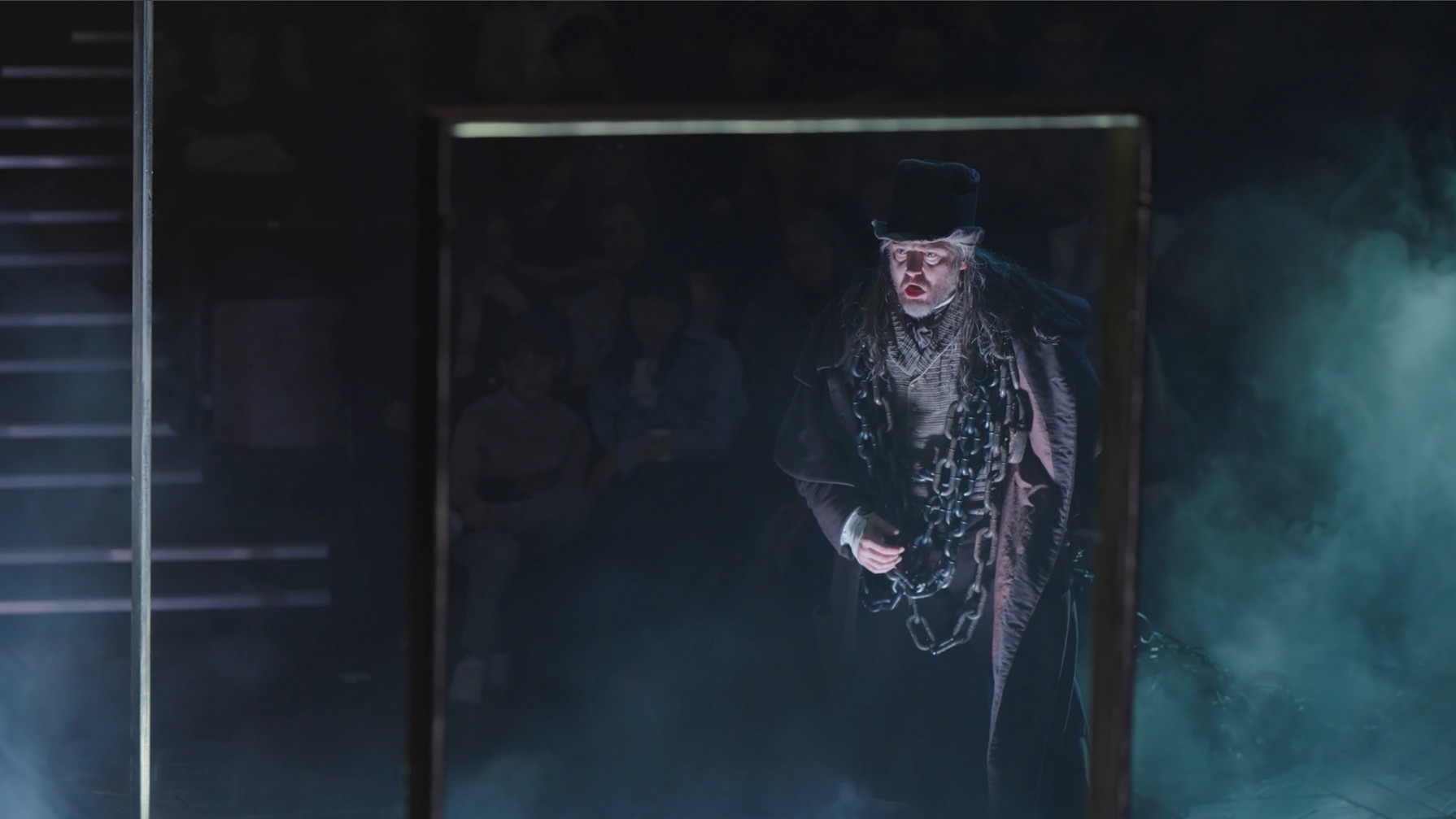 Actor Stuart Neal wears a large dark coat and a top hat. He's wrapped in a large chain and stands on a dark foggy stage.
