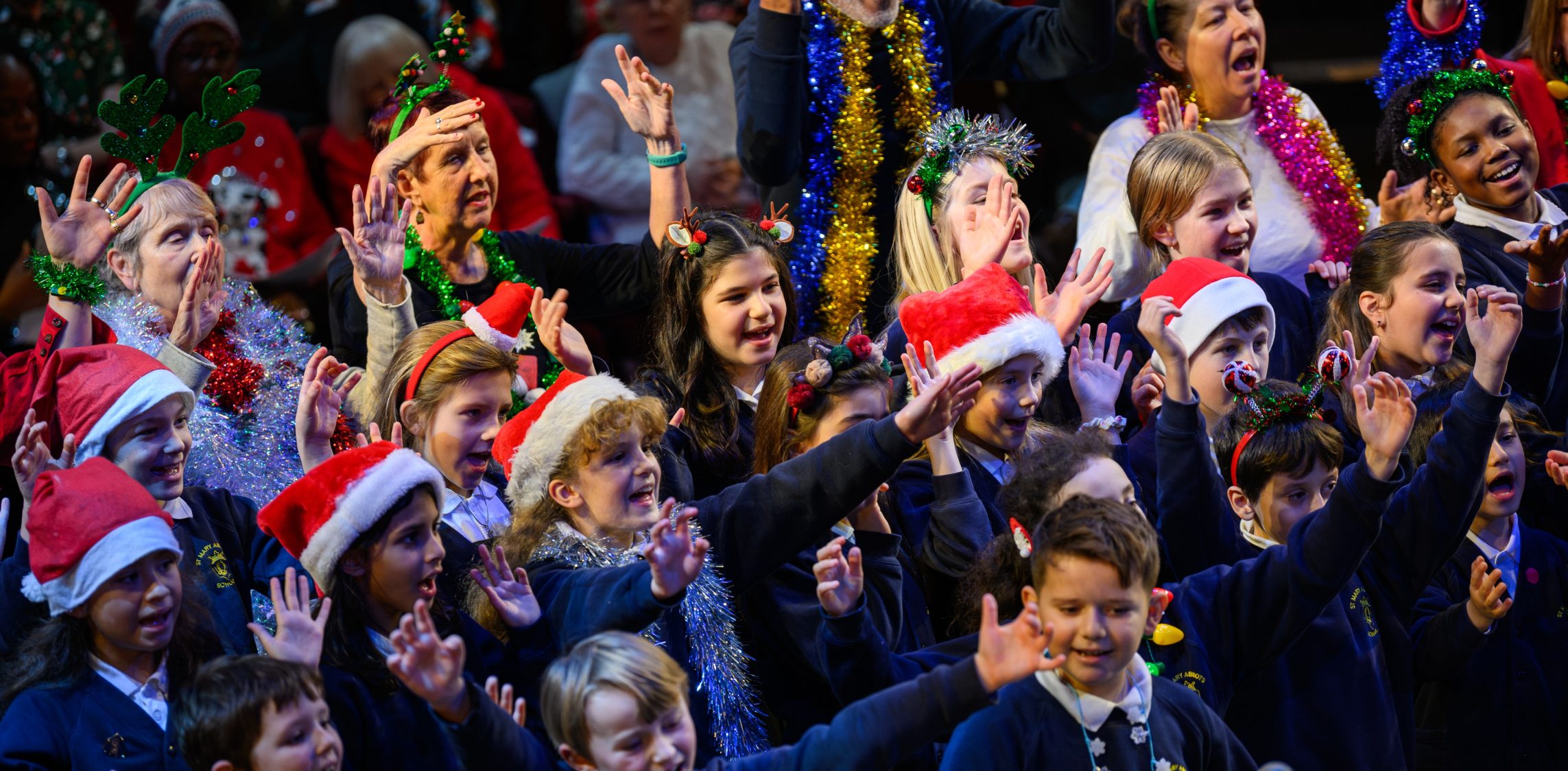 A choir is singing, made up of primary school children wearing school uniforms and Christmas accessories and older people dressed in Christmas jumpers and tinsel.