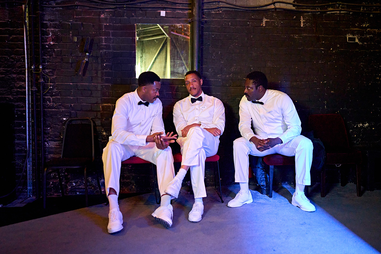 Samson Ajewole, Jaz Hutchins and Kazeem Tosin Amore sit side by side on three chairs backstage. They are chatting to each other, wearing white shirts, white trousers and black bow ties.