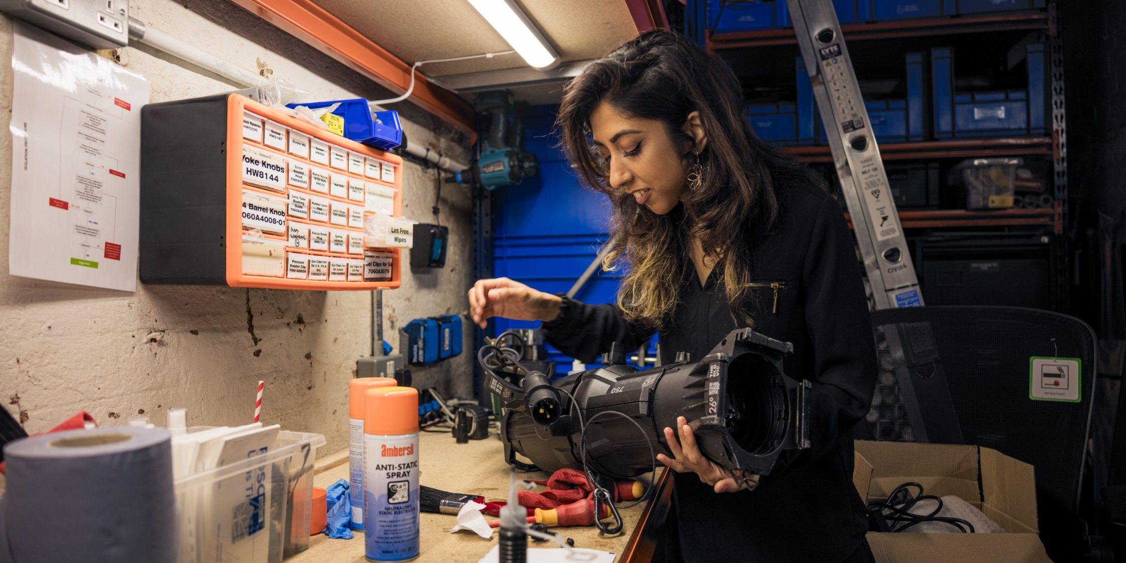 A young woman is holding onto a theatre light that is resting on a desk cluttered with technical equipment