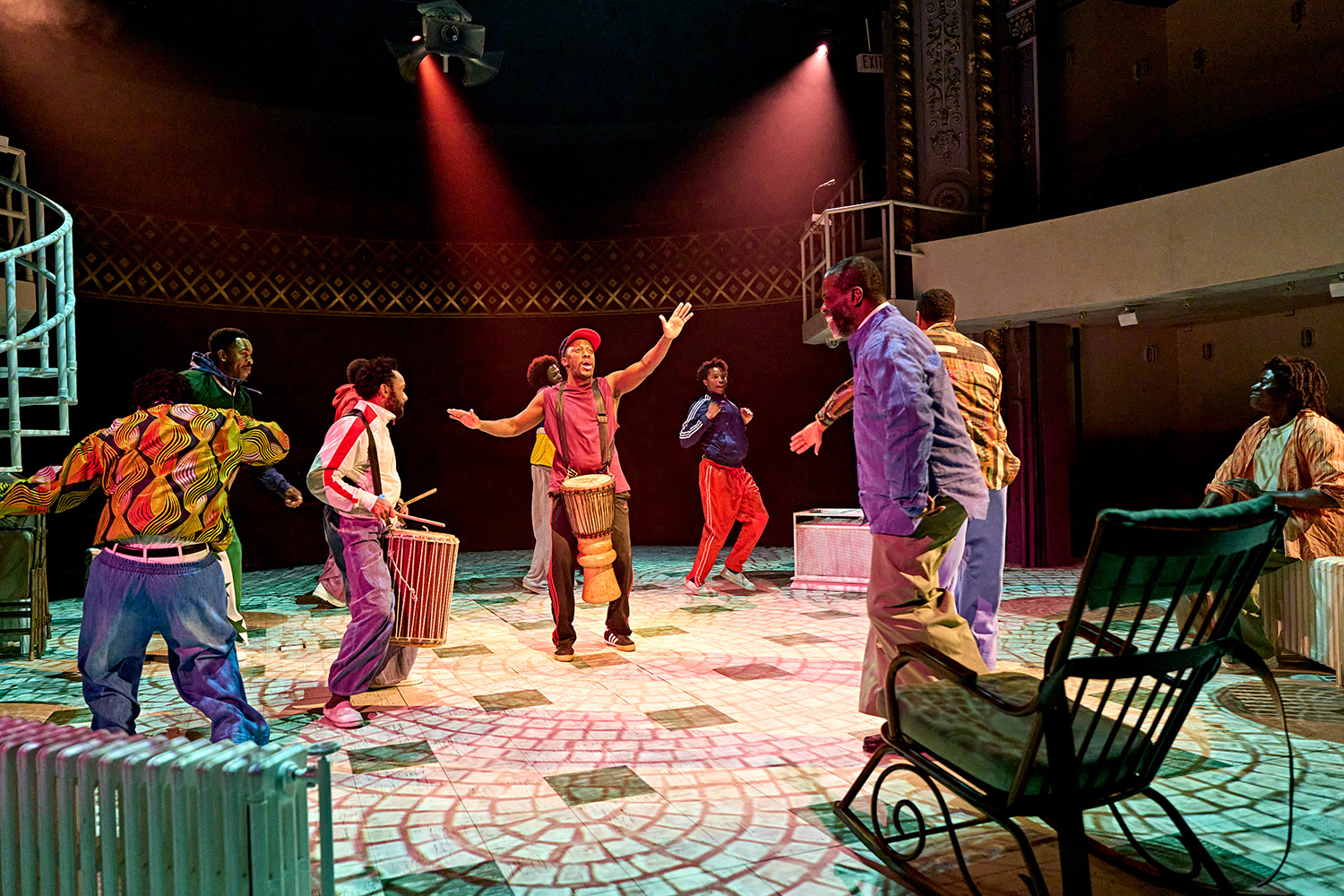 A group of actors gather with drums and dancing on a stage with a projection of cobbles. They are wearing brightly coloured shirts, baggy jeans or tracksuits.