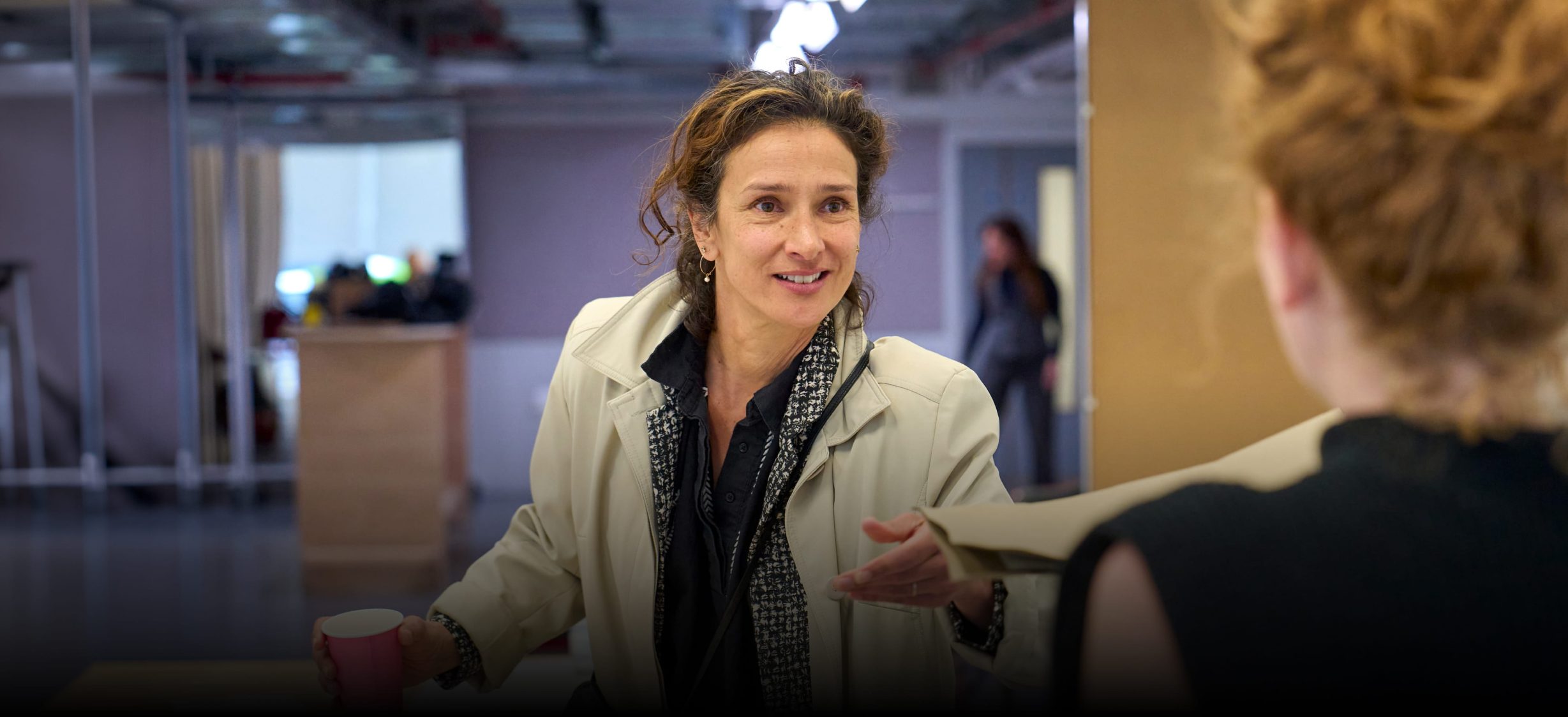 Indira Varma in rehearsals for Glengarry Glen Ross. She wears a beige Macintosh, holds a pink disposable coffee cup and reaches forward to retrieve a file from a woman standing in the foreground.