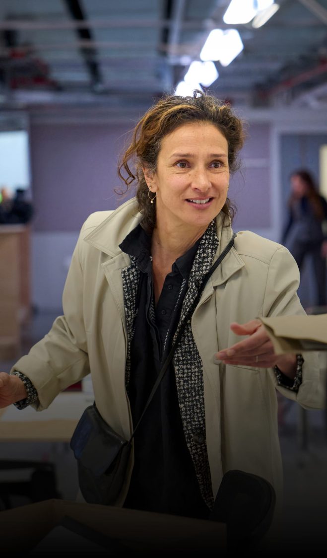 Indira Varma in rehearsals for Glengarry Glen Ross. She wears a beige Macintosh, holds a pink disposable coffee cup and reaches forward to retrieve a file from a woman standing in the foreground.