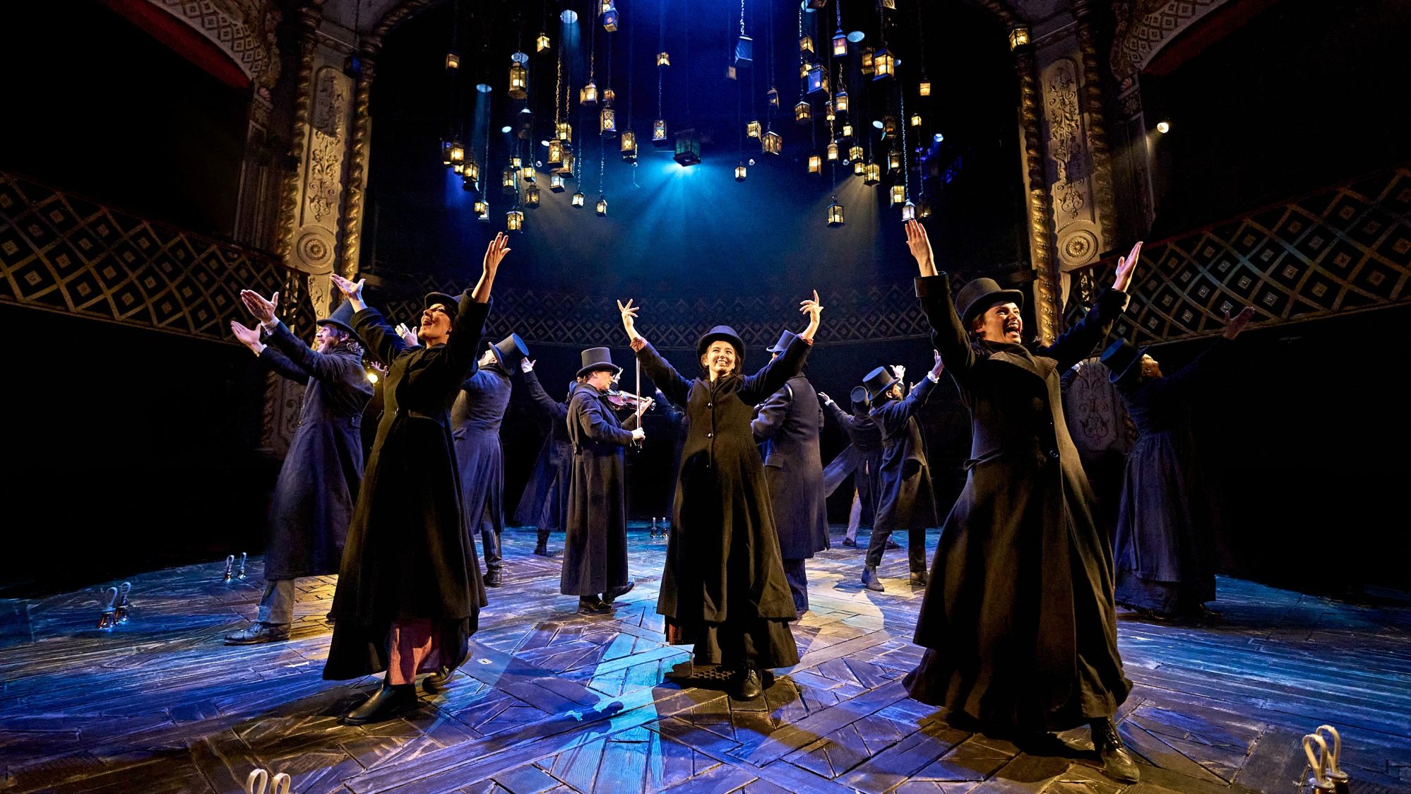 The cast of A Christmas Carol stand in a circle with their arms raised towards the audience. They are all wearing long black coats and top hats. Glowing lanterns hand from the ceiling above them.