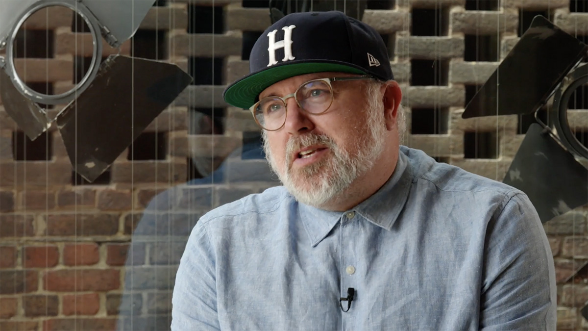 Phillip Breen looks into the middle distance. He's wearing a light blue button up shirt and a dark blue cap with an 'H' on it. He's sitting in front of a window which looks out on some hanging recycled theatre lights.