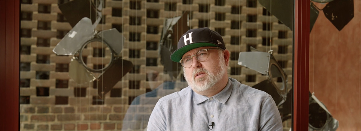 Phillip Breen looks into the middle distance. He's wearing a light blue button up shirt and a dark blue cap with an 'H' on it. He's sitting in front of a window which looks out on some hanging recycled theatre lights.