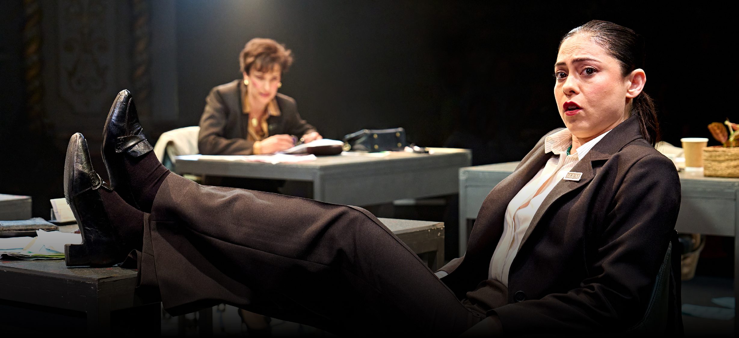Rosa Salazar leans back on a desk chair and places her feet on the desk in front of her. She wears a white shirt and black suit jacket and trousers.