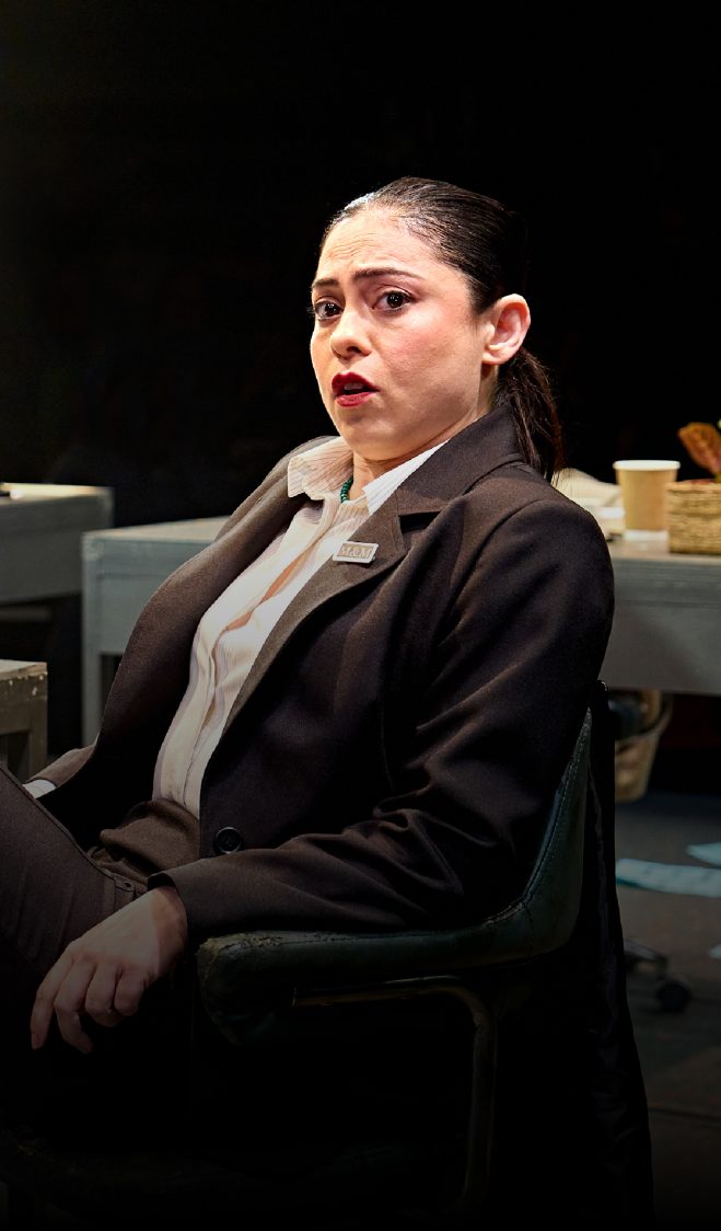 Rosa Salazar leans back on a desk chair and places her feet on the desk in front of her. She wears a white shirt and black suit jacket and trousers.