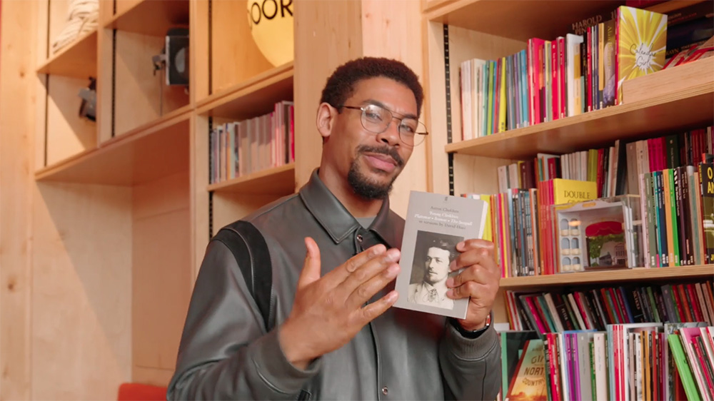 Aaron Pierre smiles confidently at the camera, holding a grey book in his hand with a black and white photo on the cover, next to a busy bookshelf.