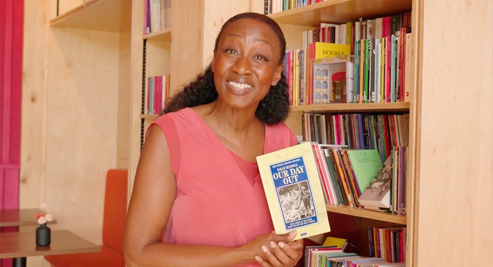Beverley Knight smiles in front of wooden shelves filled with scripts. She's holding a copy of Our Day Out and smiling to the camera