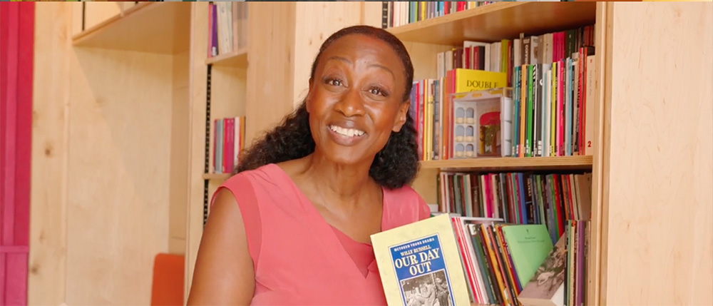 Beverley Knight smiles in front of wooden shelves filled with scripts. She's holding a copy of Our Day Out and smiling to the camera