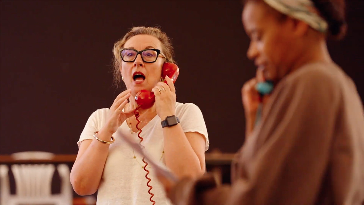 Dorothy Atkinson stands in an airy rehearsal room speaking into an old fashioned red telephone. Ayesha Antoine is out of focus in the foreground.