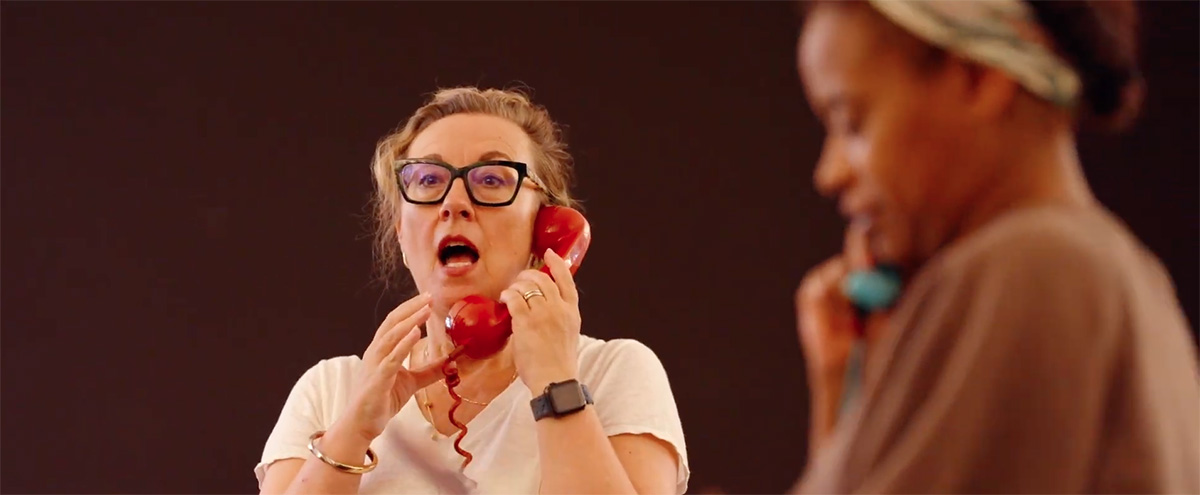 Dorothy Atkinson stands in an airy rehearsal room speaking into an old fashioned red telephone. Ayesha Antoine is out of focus in the foreground.