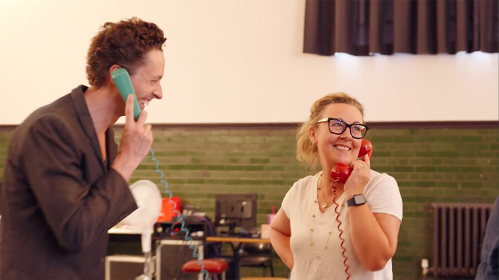 Dorothy Atkinson and Rowan Polonski stand in an airy rehearsal room holding brightly coloured old fashioned telephones up to their ears.