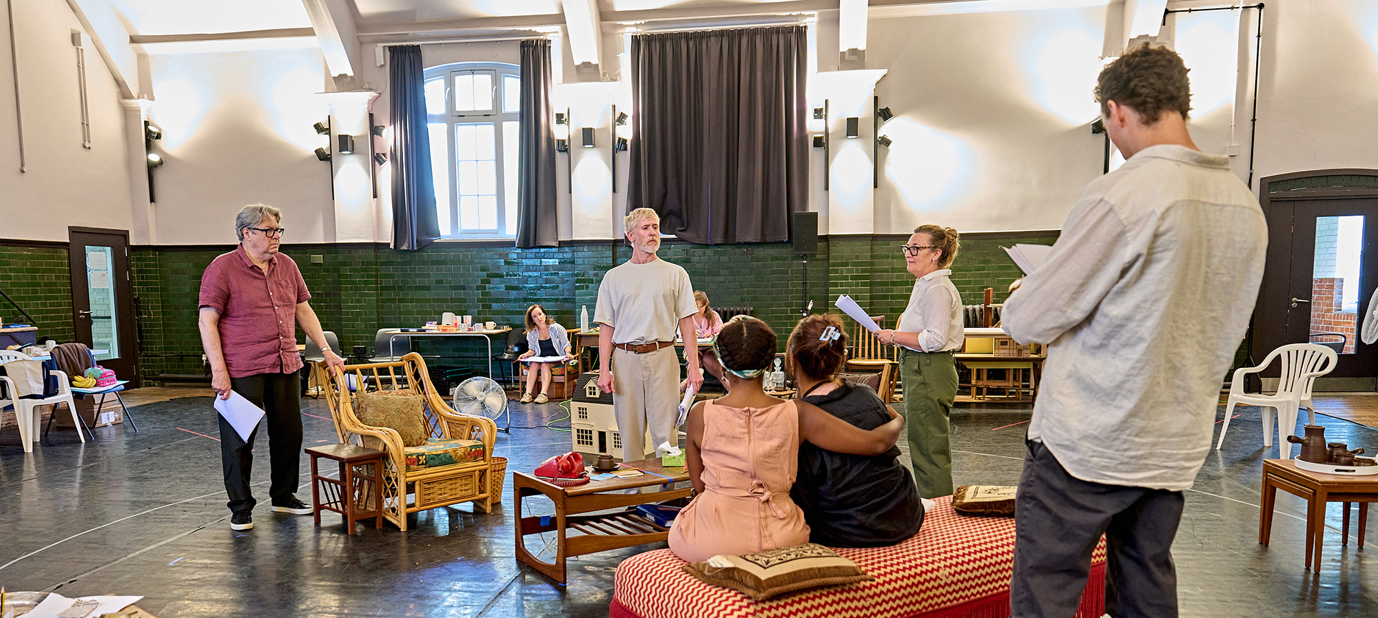The cast of How the Other Half Loves stand around the set for the show in an airy green-tiled rehearsal room. They stand with scripts in hand. There are a selection of chairs and in the middle of the room there's a table with brightly coloured old fashioned phones.