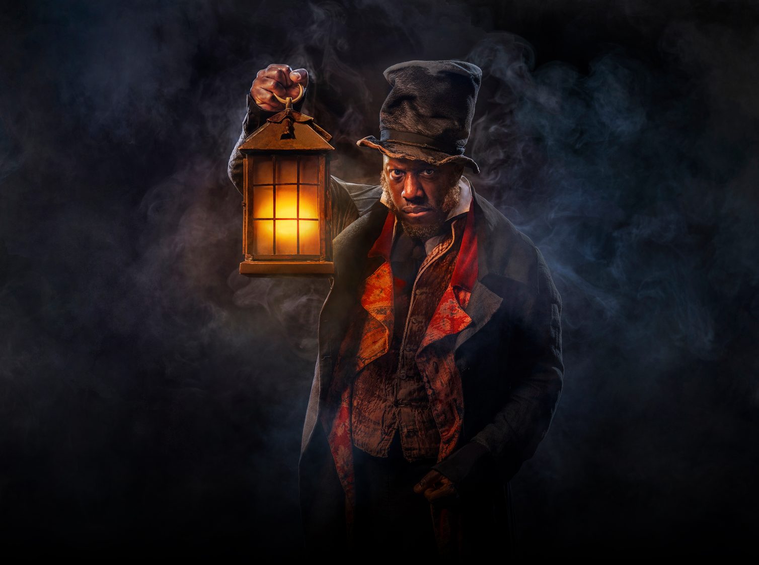 Actor Giles Terera emerges from a misty dark background. He's holding a lantern with a warm orange glow. He's wearing a crumpled top hat, a dark overcoat and a shabby waistcoat.