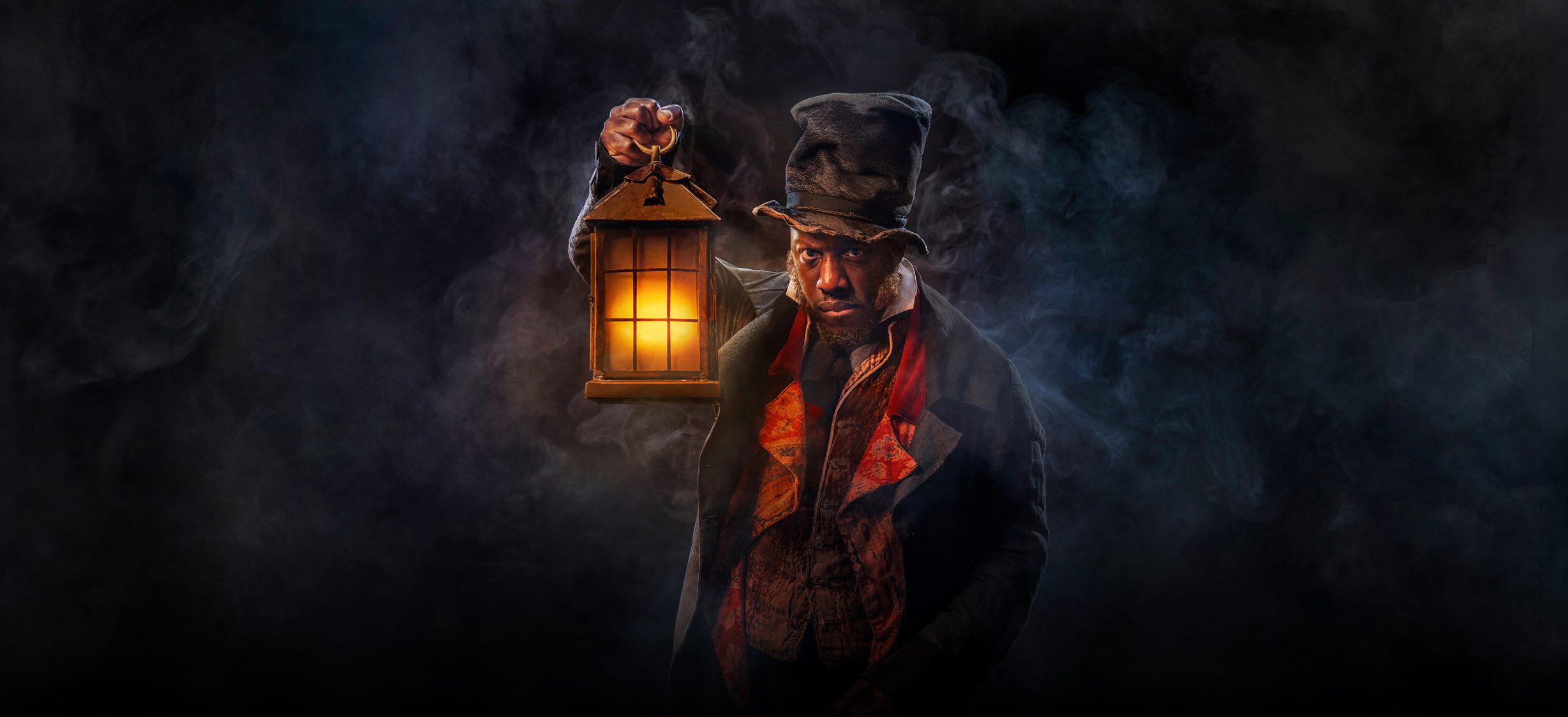 Actor Giles Terera emerges from a misty dark background. He's holding a lantern with a warm orange glow. He's wearing a crumpled top hat, a dark overcoat and a shabby waistcoat.