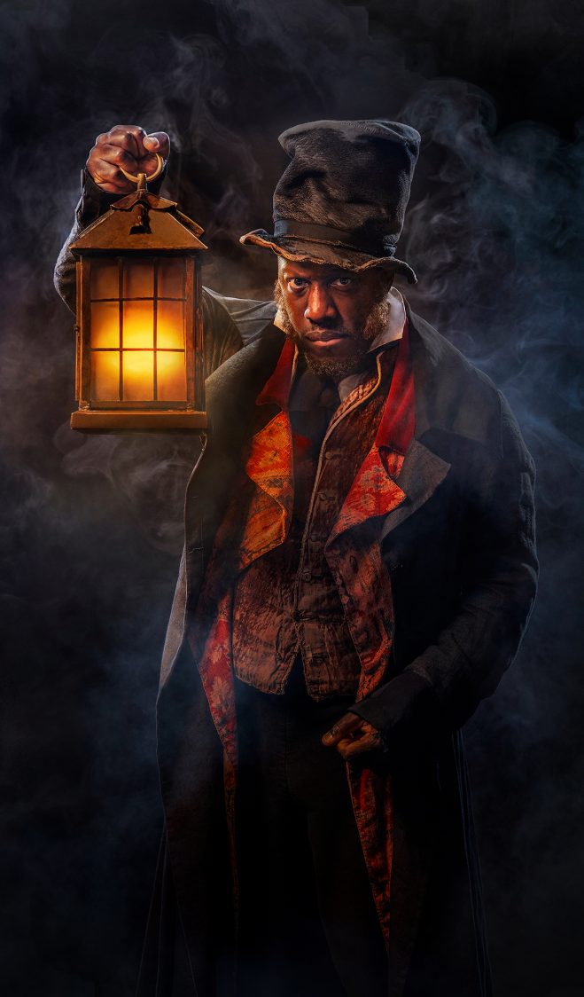 Actor Giles Terera emerges from a misty dark background. He's holding a lantern with a warm orange glow. He's wearing a crumpled top hat, a dark overcoat and a shabby waistcoat.
