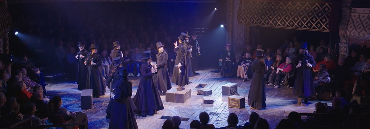 A group of actors in long dark coats and top hats stand on a round stage underneath a canopy of hanging lanterns.