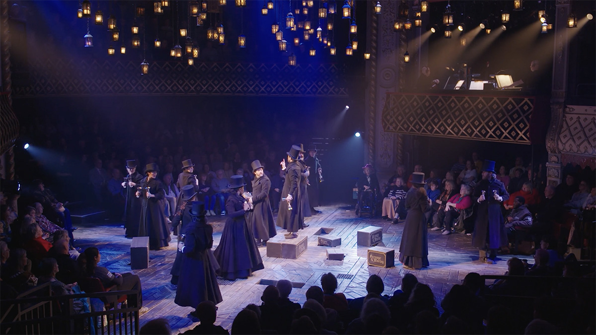 A group of actors in long dark coats and top hats stand on a round stage underneath a canopy of hanging lanterns.