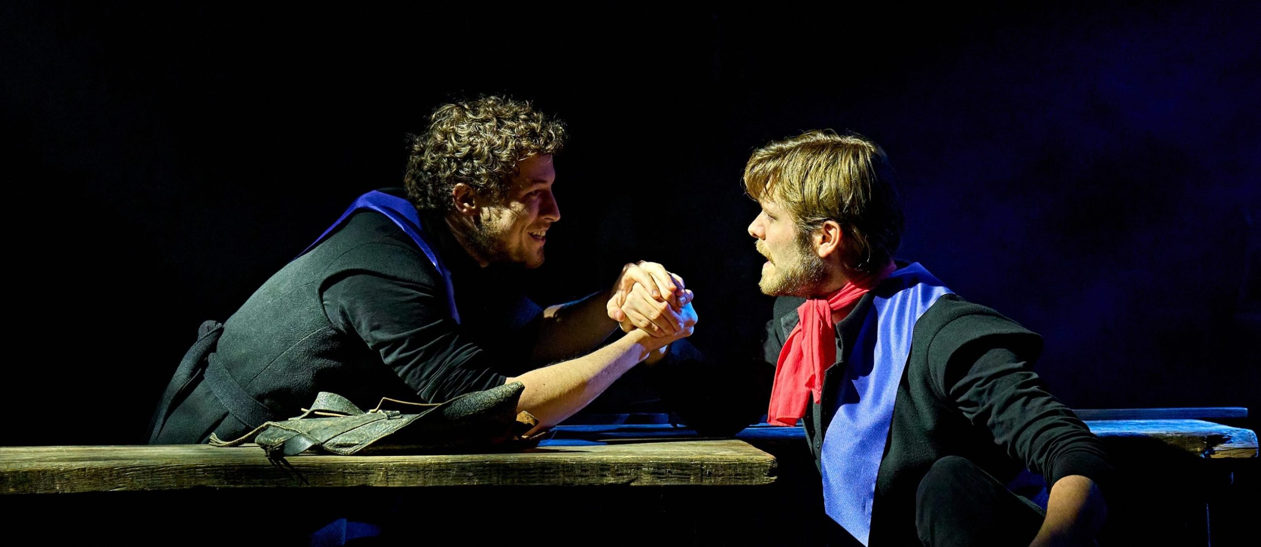 Actors Alex James-Hatton and Shane O'Riordan clasp hands at a wooden table, both wearing black costumes with red and blue accents.