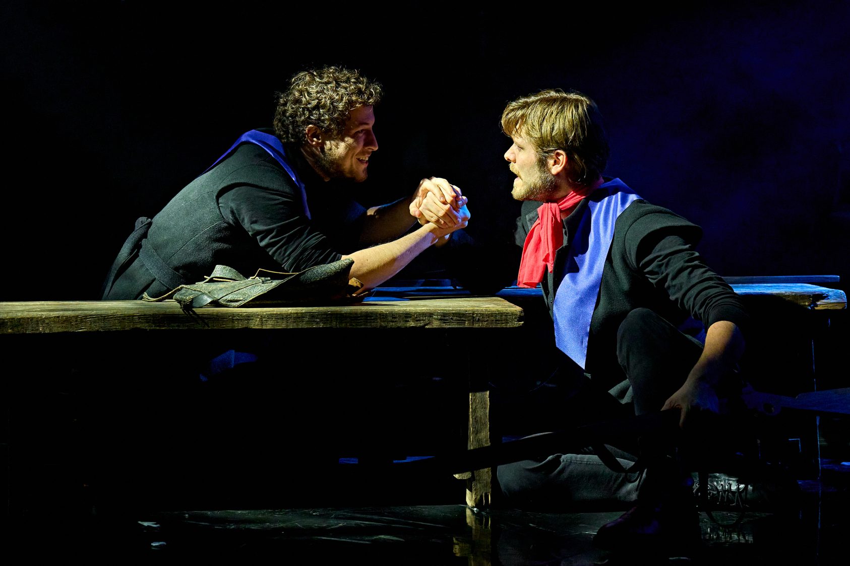 Actors Alex James-Hatton and Shane O'Riordan clasp hands at a wooden table, both wearing black costumes with red and blue accents.