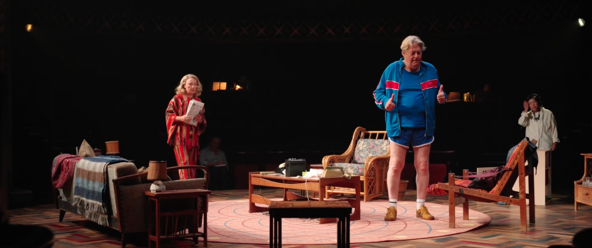 On a dark stage with a domestic set actor Roger Allam stands in a blue tracksuit with blue shorts. Actor Dorothy Atkinson stands behind him wearing an orange striped dress and matching headband, holding a newspaper.