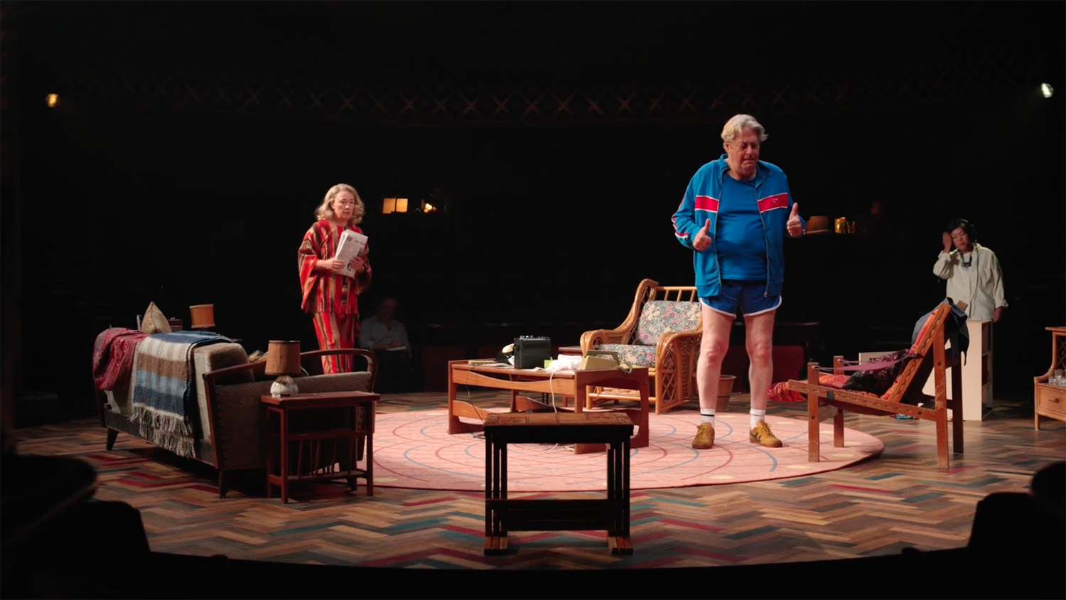 On a dark stage with a domestic set actor Roger Allam stands in a blue tracksuit with blue shorts. Actor Dorothy Atkinson stands behind him wearing an orange striped dress and matching headband, holding a newspaper.