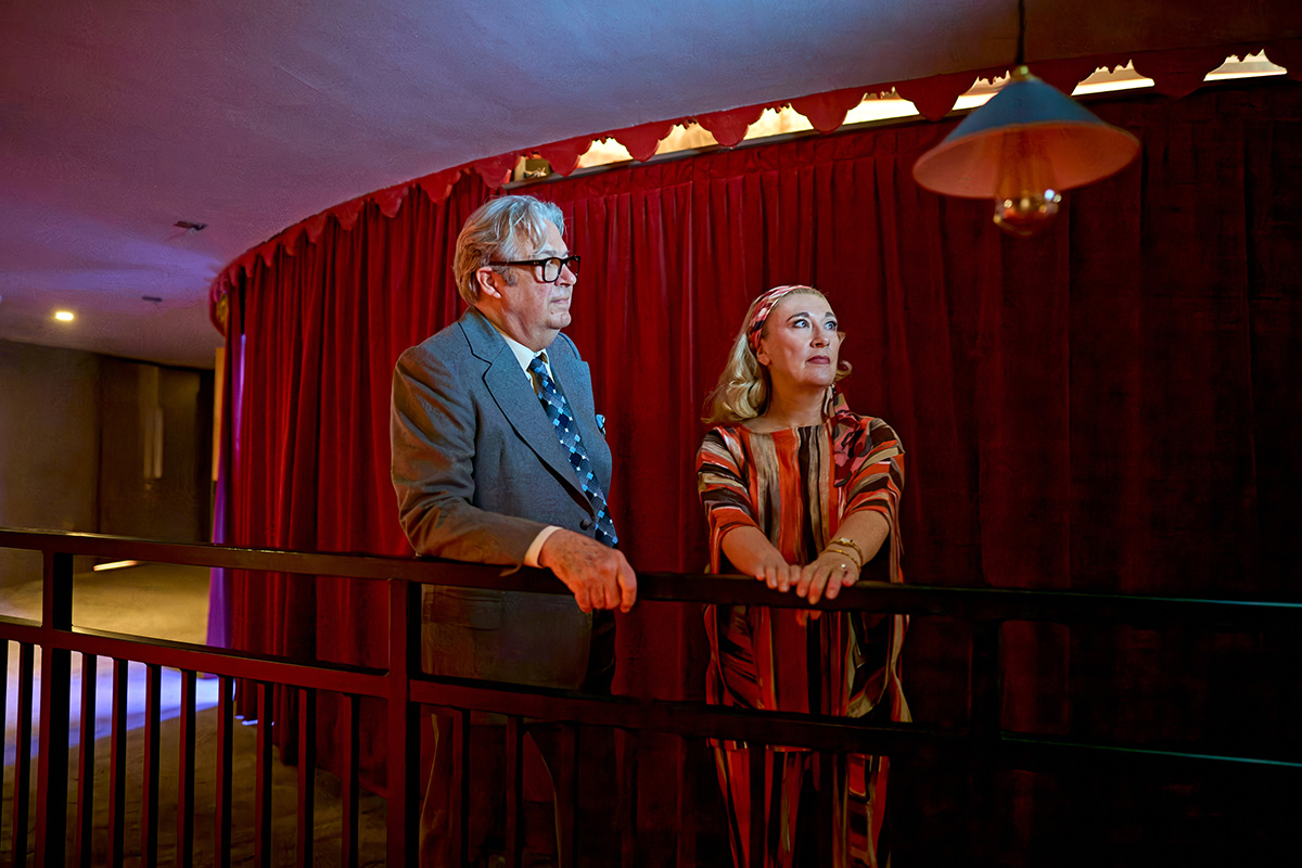 Roger Allam and Dorothy Atkinson stand in the wings waiting to go on stage. Roger wears a grey suit with a blue tie and leans on a handrail. Dorothy wears a red striped 70s style dress and matching headband.