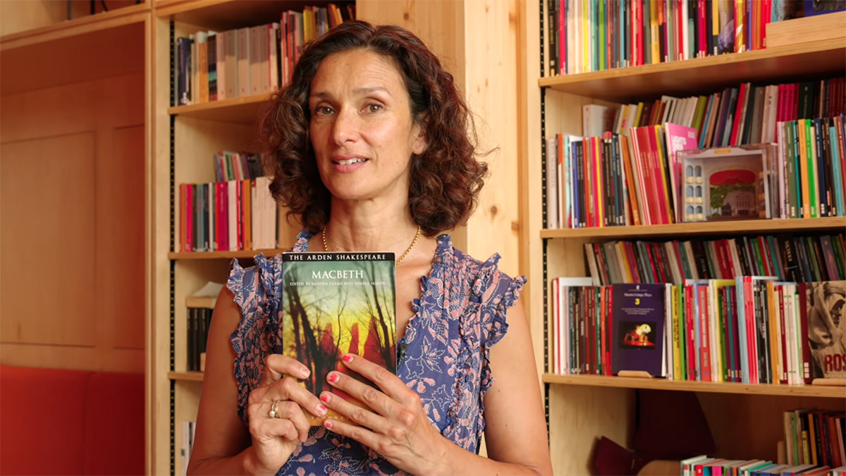 Indira Varma stands in front of a floor to ceiling bookshelf wearing a summer dress. She's holding a copy of Macbeth in front of her.