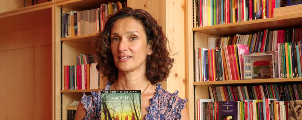 Indira Varma stands in front of a floor to ceiling bookshelf wearing a summer dress. She's holding a copy of Macbeth in front of her.