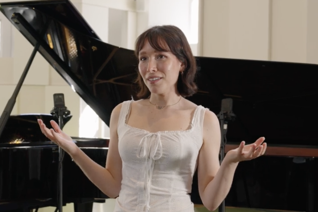 Actor Liv Andrusier has mid-length brown hair and wears a white blouse. She holds up her hands mid-conversation as she stands in front of a grand piano.