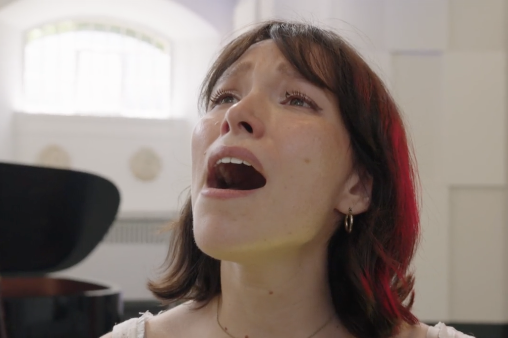 A close up of actor Liv Andrusier, who has mid-length brown hair. She is singing, looking skyward with an emotional expression, and a grand piano in the background.