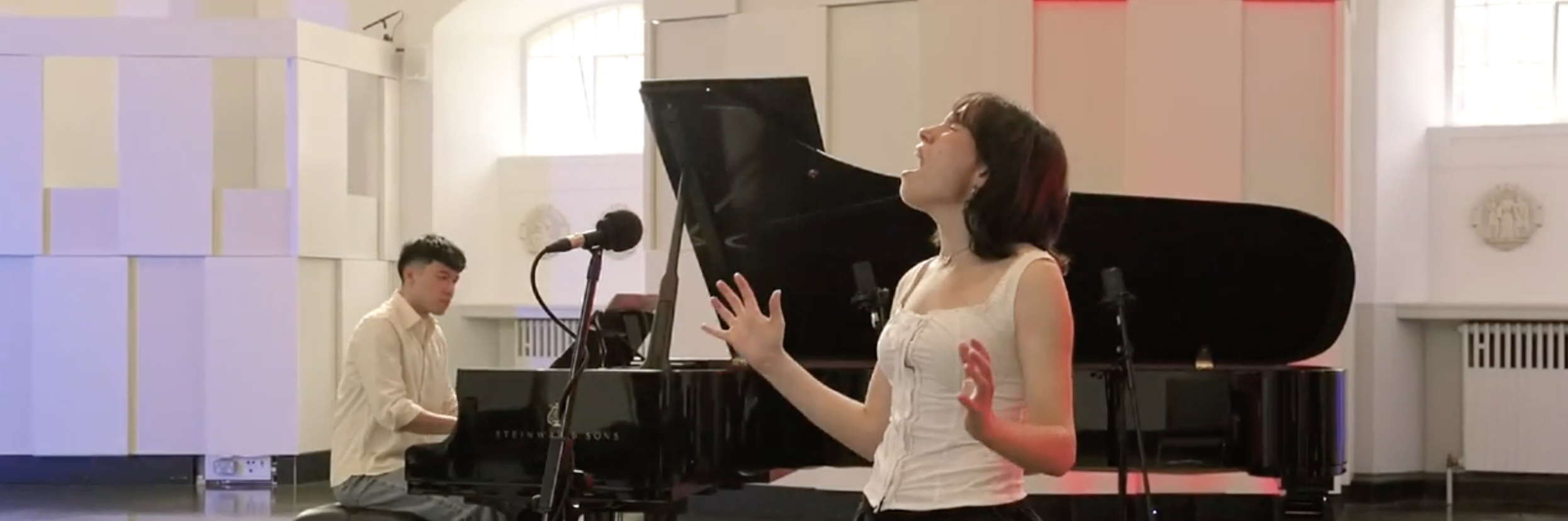 Liv Andrusier wears a white vest and black trousers as she sings passionately into a microphone. Behind her is Chris Poon playing a grand piano.