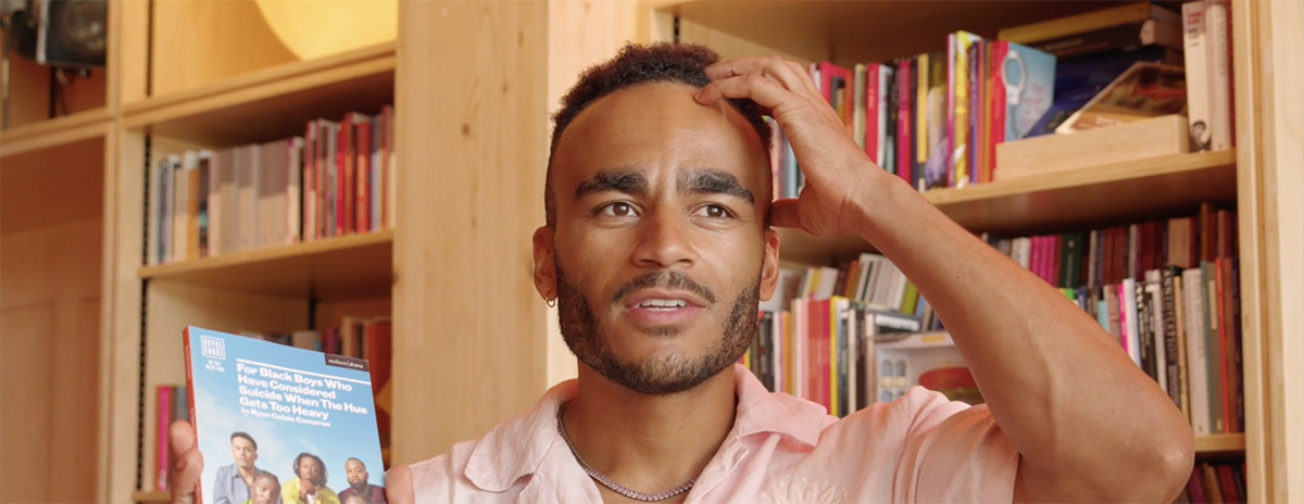 Munya Chawawa holds up a book in one hand and raises the other to his head. He's standing in front of a floor to ceiling bookshelf