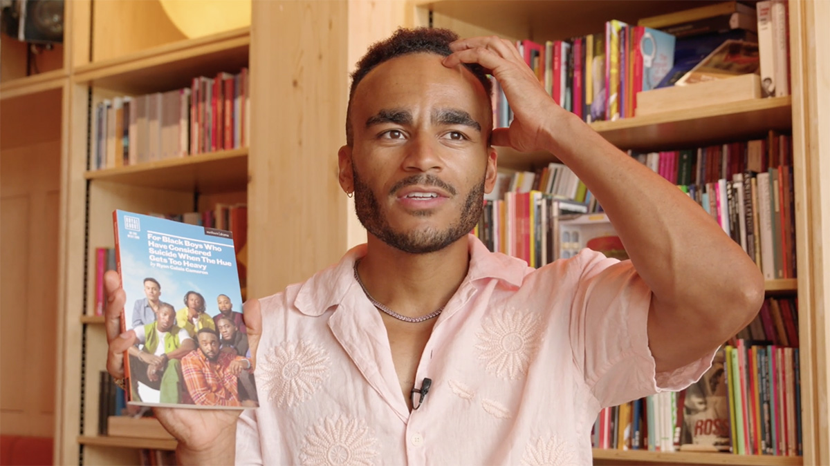 Munya Chawawa holds up a book in one hand and raises the other to his head. He's standing in front of a floor to ceiling bookshelf