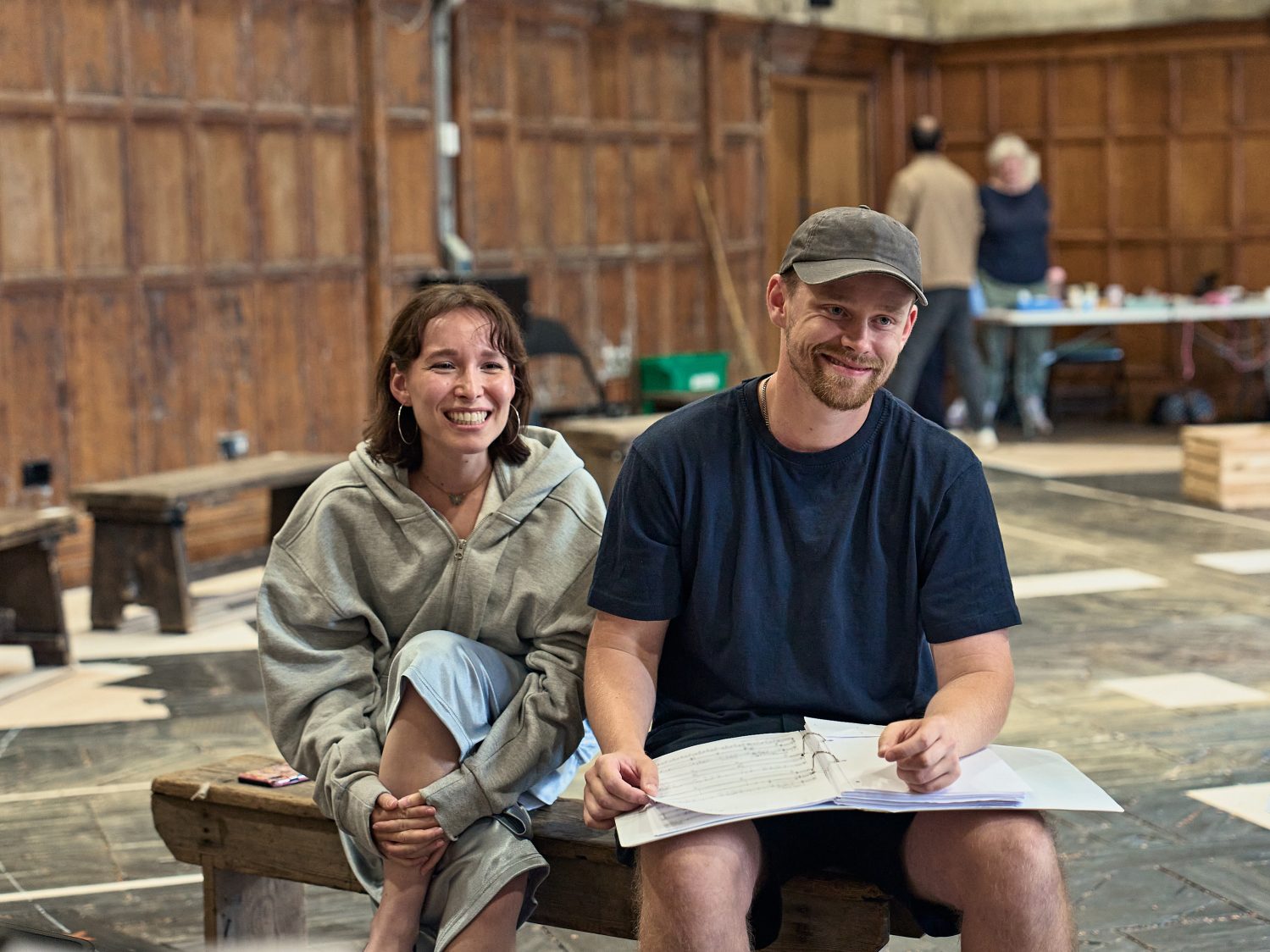 Actors Liv Andrusier and Barney Wilkinson smile as they sit together in the rehearsal room. Liv is cross-legged, Barney holds an open musical score.