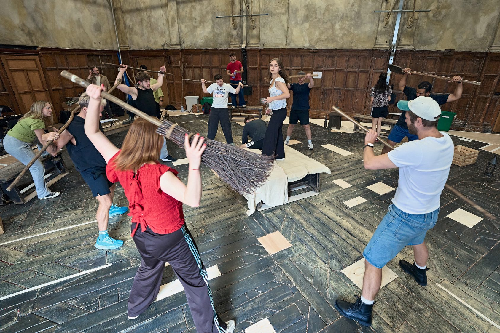 The cast of Martin Guerre rehearse, encircling a lone central figure. They hold wooden farm tools and brooms above their heads.