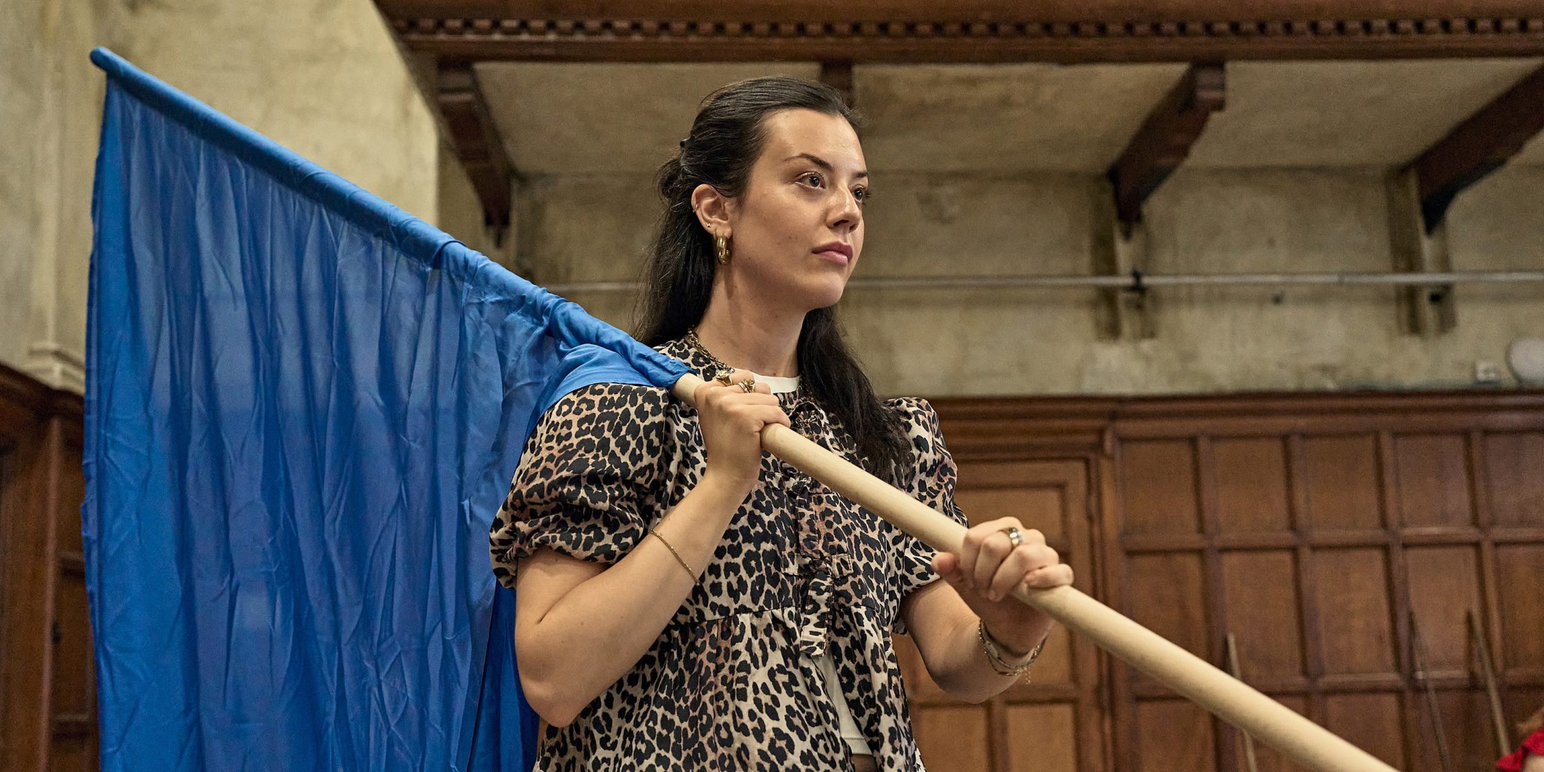 Actor Grace Galloway stands in the Martin Guerre rehearsal room wearing a leopard blouse and hoisting a blue flag over her shoulder.