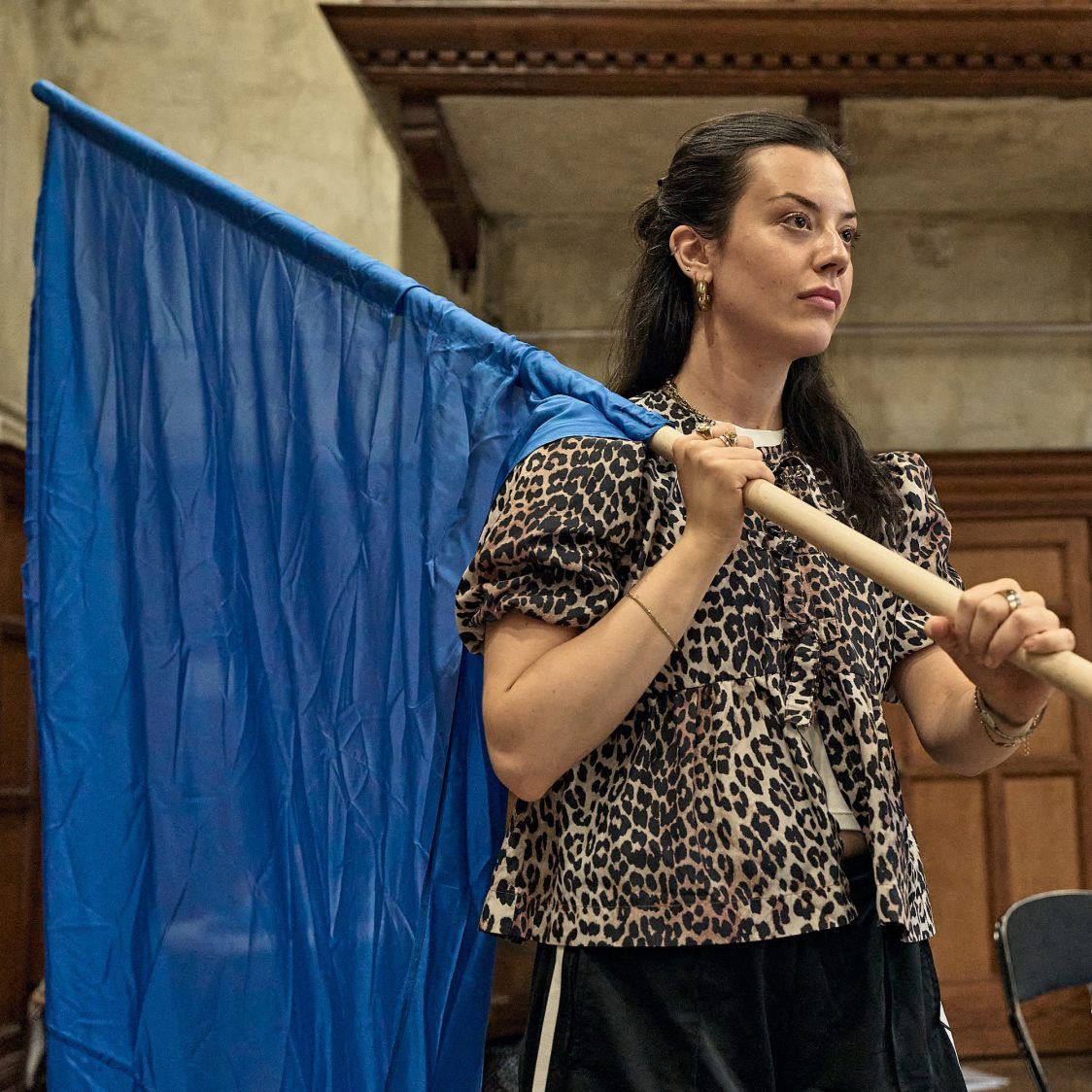 Actor Grace Galloway stands in the Martin Guerre rehearsal room wearing a leopard blouse and hoisting a blue flag over her shoulder.
