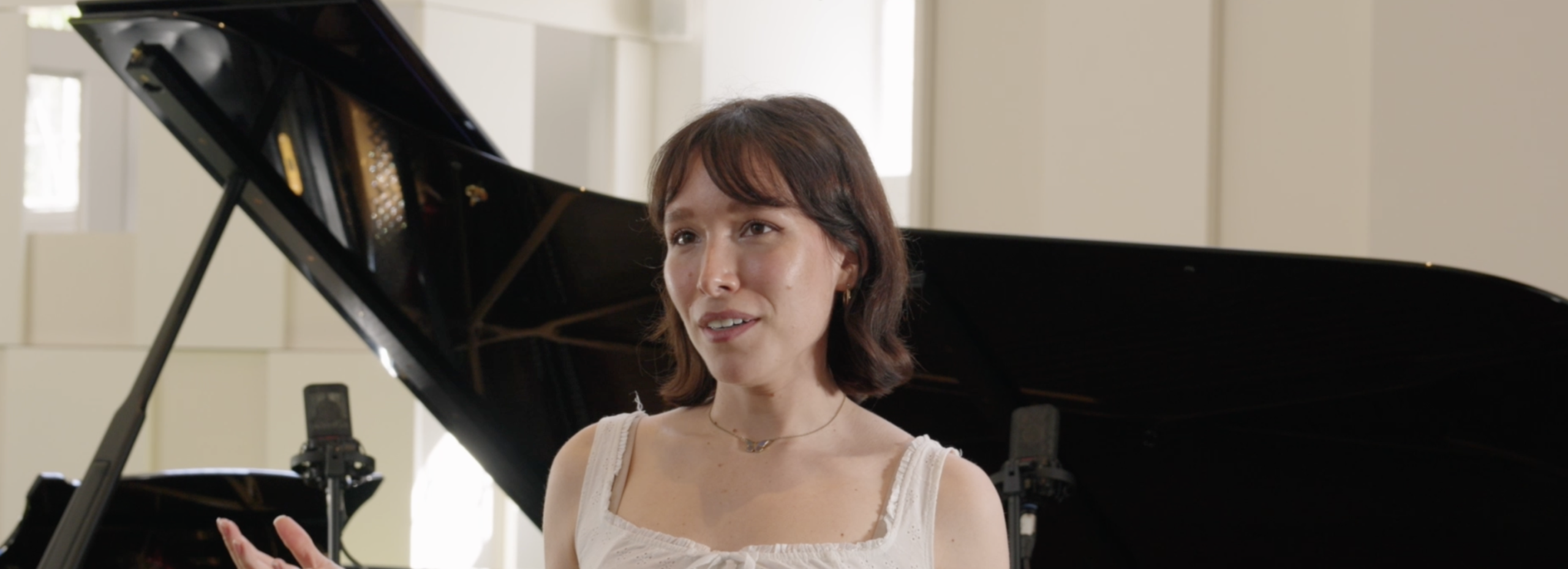 Actor Liv Andrusier has mid-length brown hair and wears a white blouse. She holds up her hands mid-conversation as she stands in front of a grand piano.