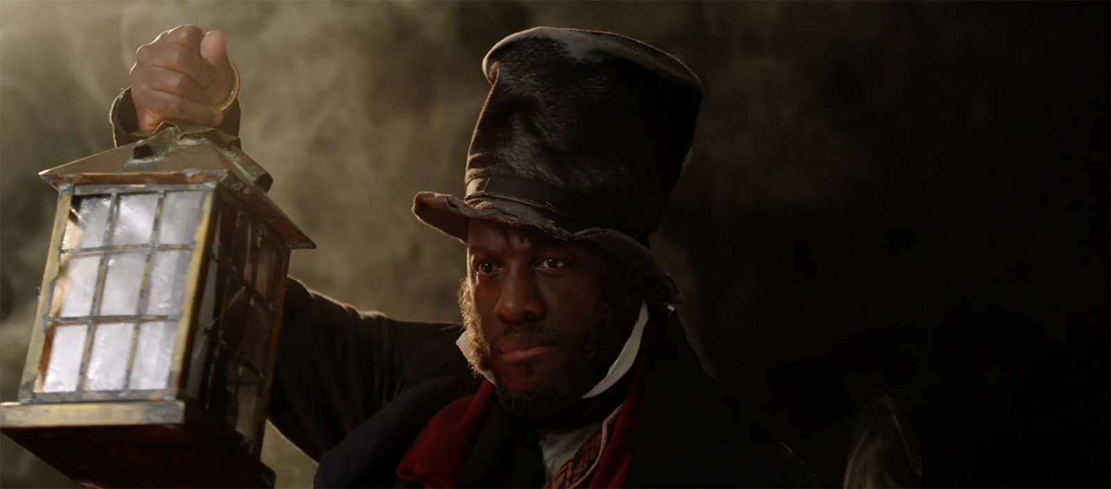 Actor Giles Terera emerges from a misty dark background. He's holding a lantern with a warm orange glow. He's wearing a crumpled top hat, a dark overcoat and a shabby waistcoat.